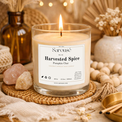 A lit Sarouse candle in a clear glass jar sits on a woven coaster surrounded by warm autumn props, including crystals, dried flowers, wooden beads, and a matchbox. The label reads 'Harvested Spice Pumpkin Chai'.