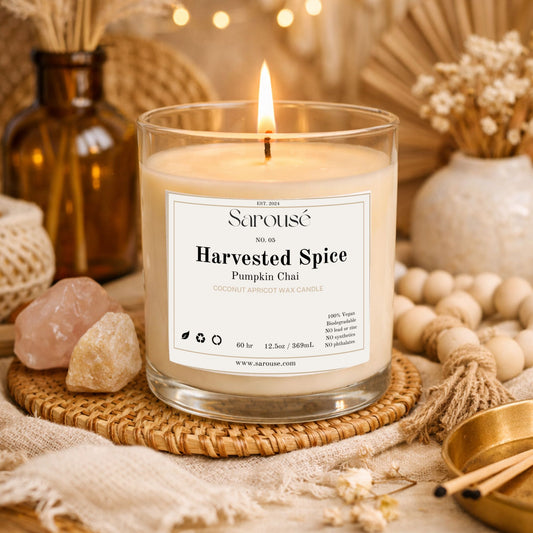 A lit Sarouse candle in a clear glass jar sits on a woven coaster surrounded by warm autumn props, including crystals, dried flowers, wooden beads, and a matchbox. The label reads 'Harvested Spice Pumpkin Chai'.